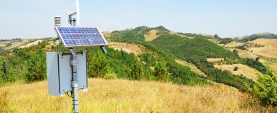 A solar-powered weather station stands in a grassy field with rolling hills and trees in the background under a clear blue sky.