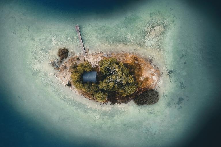 Aerial view of a small island with a single building, trees, a dock, and shallow water surrounding it.