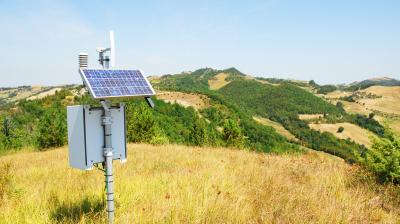 A solar-powered weather station stands in a grassy field with rolling hills and trees in the background under a clear blue sky.