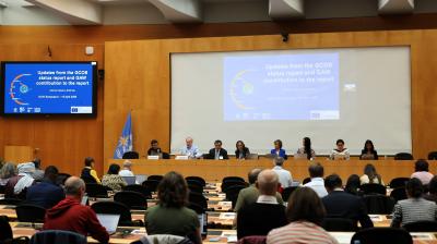 A panel of eight people sits at a conference table in front of an audience, with presentation slides displayed on large screens behind them in a modern meeting room.