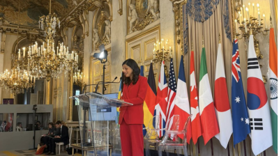 A woman in a red suit speaks at a clear podium in an ornate room with chandeliers and multiple national flags displayed behind her.