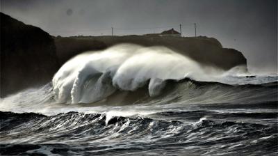 Large ocean waves crash near a rocky coast under a cloudy sky, with a silhouette of a cliff and buildings in the background.