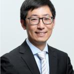A man with short black hair and glasses wearing a black suit, white shirt, and striped tie smiles in front of a plain background.