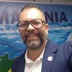 A man with glasses and a beard smiles at the camera. He is wearing a blue jacket and white shirt, standing in front of a blue and green banner with text.