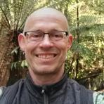 A bald man with glasses and a backpack smiles at the camera, standing outdoors in a lush, green wooded area.