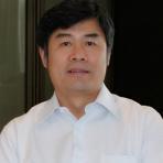 A man with short black hair wearing a white button-up shirt is looking at the camera with a neutral expression. The background includes a partially visible window or glass panel.