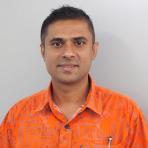 A man wearing a bright orange shirt with geometric patterns stands against a plain light grey background, looking directly at the camera.
