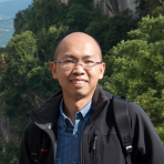 A person with glasses and a backpack smiles at the camera, standing outdoors with trees and hills in the background.