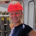 A person wearing a red hard hat and blue shirt smiles while standing on a ship or industrial vessel.
