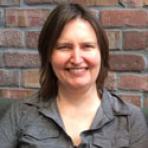 A person with shoulder-length brown hair and a grey shirt smiles while sitting in front of a brick wall.