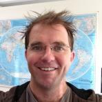 Person with glasses, smiling, standing in front of a world map on a wall.