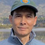 A person wearing a green cap and gray jacket poses outdoors with a mountainous landscape in the background.
