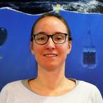 A person wearing glasses with long hair is smiling while standing in front of a display that includes images of underwater equipment and ocean scenes.