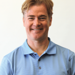 A man with light brown hair is smiling, wearing a light blue polo shirt, standing against a plain white background.