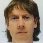 A person with medium-length, light brown hair, and blue eyes is shown in a close-up, straight-faced portrait against a plain background.