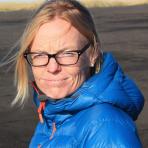 A person with blonde hair, wearing glasses and a blue puffer jacket, stands outdoors with a clear sky in the background.