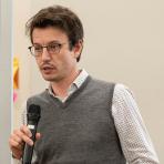 A man with short brown hair, glasses, and a gray sleeveless sweater over a white patterned shirt is holding a microphone.