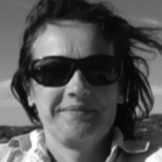 Black and white photo of a person wearing sunglasses, with windblown hair, and a slight smile, outdoors.