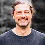 A man with long gray hair and a beard, wearing a dark sweater, smiles at the camera in a blurred outdoor setting.