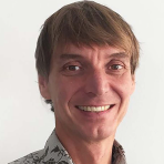 A person with brown hair is smiling, wearing a patterned shirt, against a plain white background.