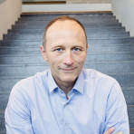 A person in a light blue shirt sits on stairs, facing the camera, with a neutral expression.