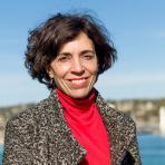 A woman in a red top and gray coat stands outdoors with a waterfront and bridge in the background under a clear blue sky.
