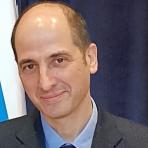 A person with short hair is wearing a suit and tie, standing in front of a dark curtain with a hint of a blue and white patterned flag visible.