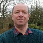 A man in a green sweater and plaid shirt stands outdoors in a garden with bare trees in the background.