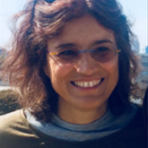 A person with shoulder-length brown hair and glasses smiles while wearing a green shirt and sunglasses on their head.
