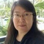 Person with long dark hair and glasses standing outdoors with palm trees in the background.
