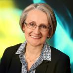A woman with short gray hair and glasses, wearing a dark blazer over a patterned shirt, smiles at the camera against a blue and yellow abstract background.