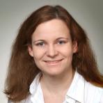 A woman with light skin, blue eyes, and shoulder-length brown hair is smiling at the camera, wearing a white collared shirt against a neutral background.
