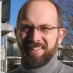 A man with glasses, a beard, and a receding hairline, wearing a gray turtleneck sweater, stands outdoors with buildings and leafless trees in the background.