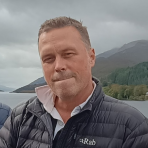 A man in a black Rab jacket stands outdoors near a body of water with mountains and trees in the background under a cloudy sky.