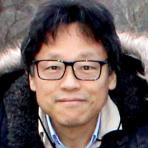 A person with short black hair and glasses is wearing a jacket with a fur-lined hood, standing outdoors.