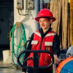 Person wearing a red hard hat and safety vest stands on an industrial worksite, holding equipment. Hoses and machinery are visible in the background.