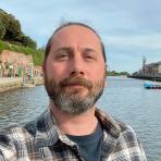 A man with a beard and plaid shirt stands by a waterfront with buildings, greenery, and boats visible in the background under a partly cloudy sky.