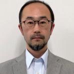 A man with glasses, a trimmed beard, and short hair is wearing a light gray suit jacket and a white shirt, standing against a plain white background.
