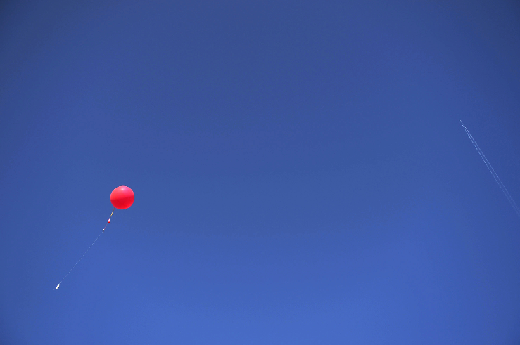 A red balloon floats in a clear blue sky with a faint trail of an airplane visible on the right.