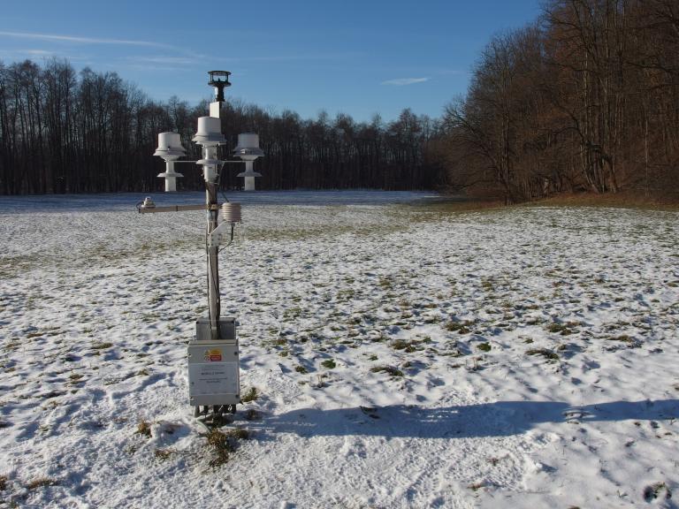 Weather station in a snow-dusted open field, with bare trees in the background under a clear blue sky.