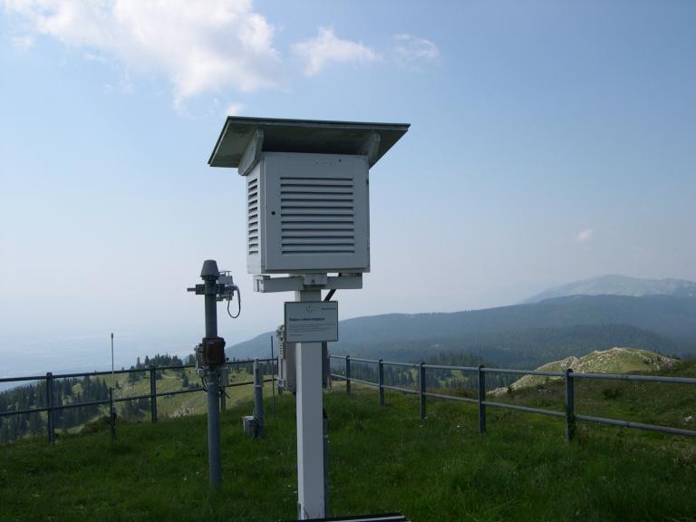 A weather station on a grassy hilltop with mountains in the background under a cloudy sky.