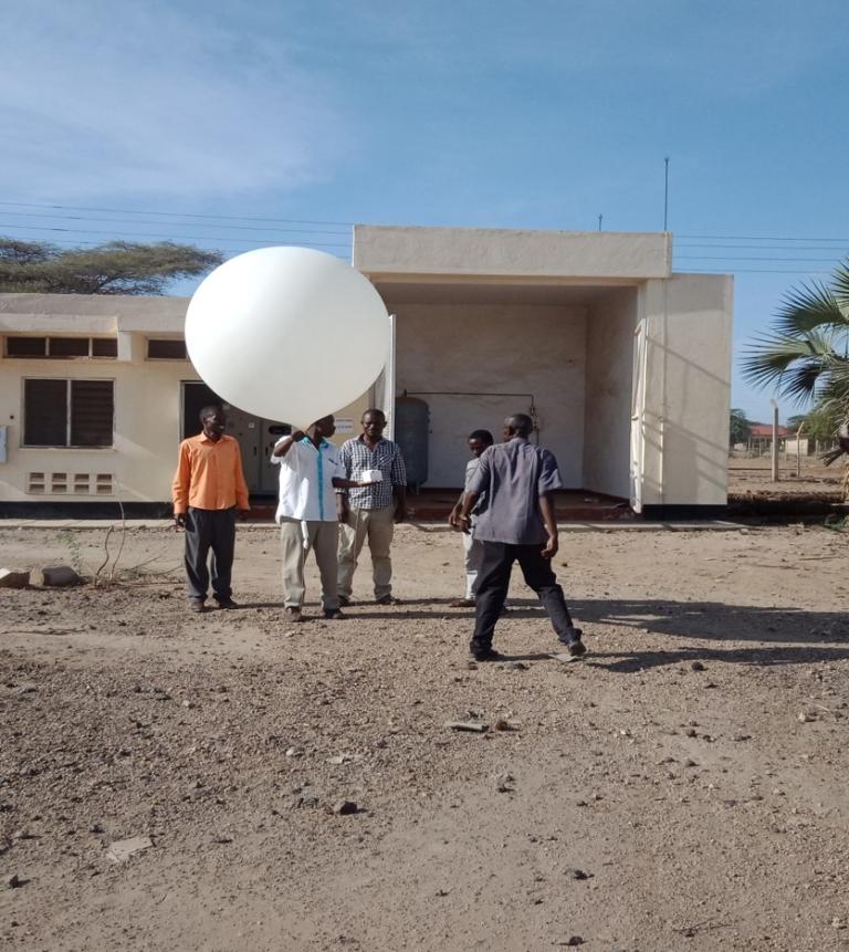 A group of people prepares to launch a large white weather balloon outside a small building on a sunny day.