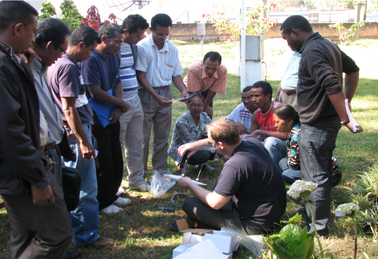 A group of people gathers outdoors around a person crouching, who is showing them a plant sample. Some hold notebooks. They appear to be engaged in a discussion or demonstration.