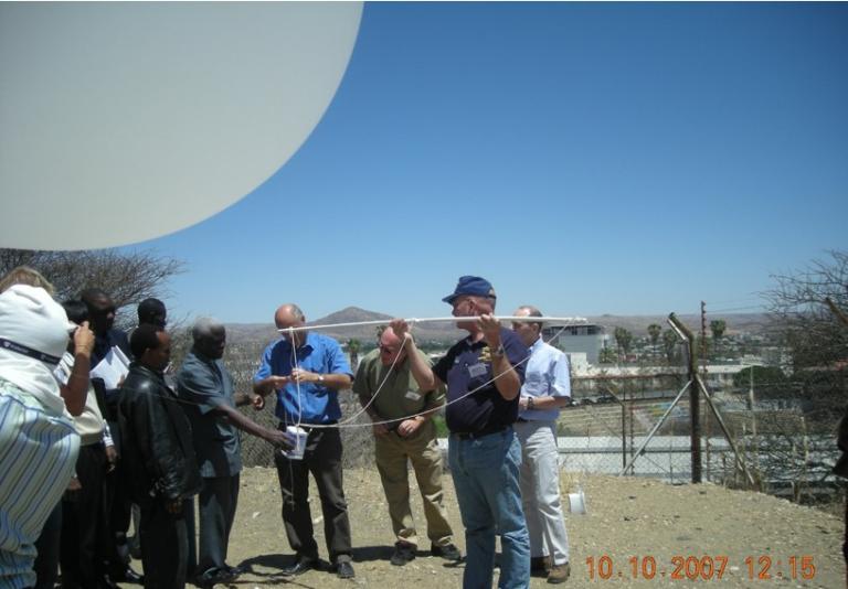 A group of people gathers outdoors to launch a weather balloon into the clear sky, equipped with instruments. Date stamp: 10.10.2007.