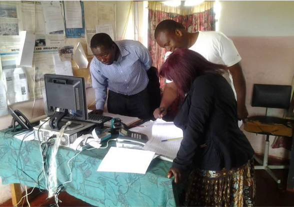 Three people in an office setting are gathered around a desk working on paperwork, with a computer and other office equipment visible.