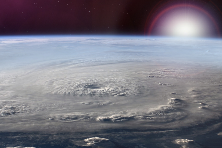 A large hurricane viewed from space with distinct spiral cloud patterns and a visible eye, set against the Earth's horizon and the sun peeking over the edge.